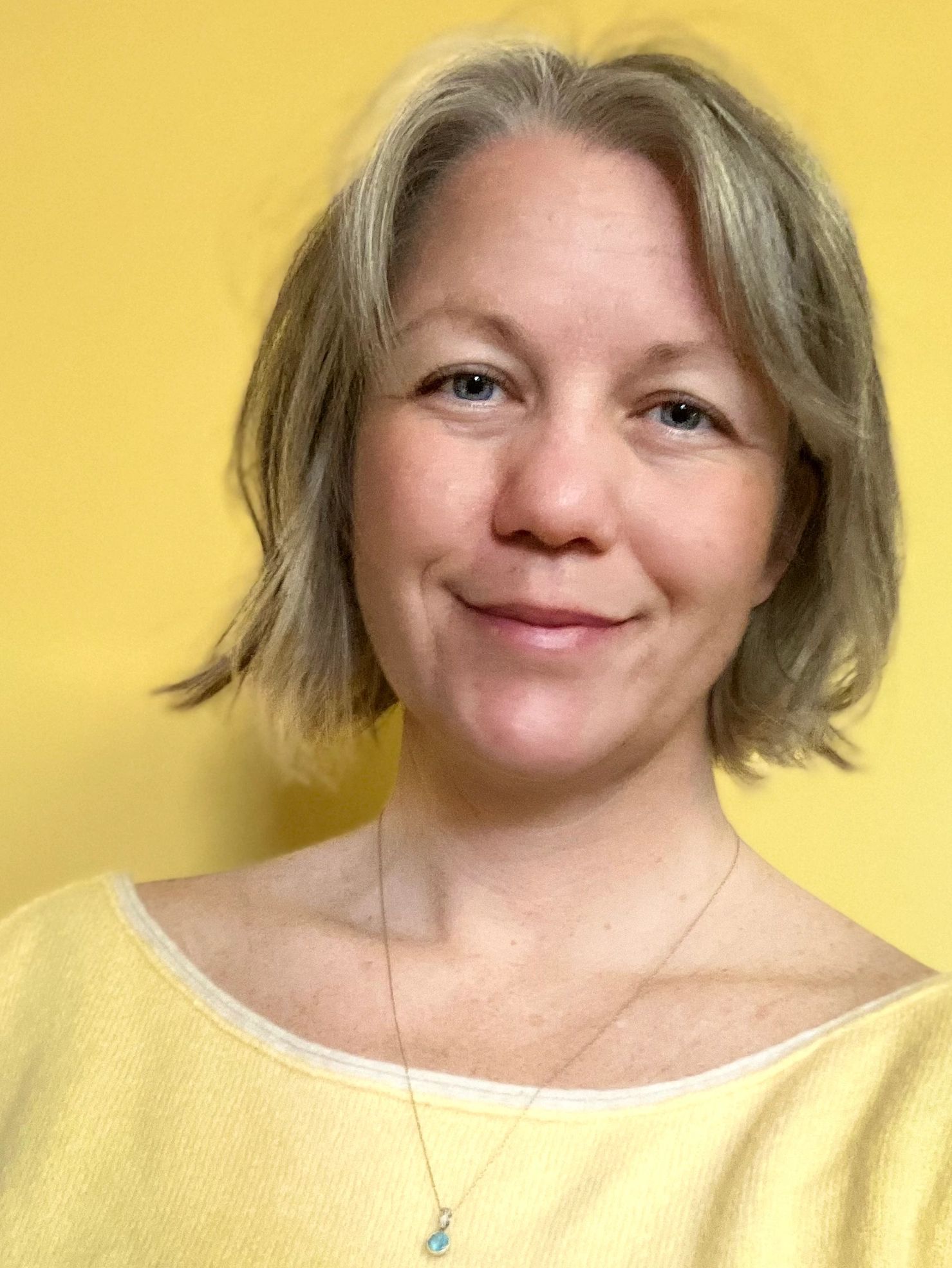 Smiling woman with short blonde hair wearing a yellow top against a yellow background.