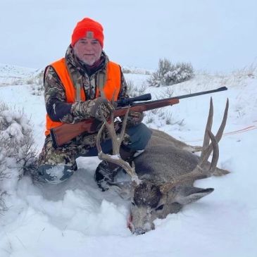 Hunter posing with a rifle and a large deer in the snow.