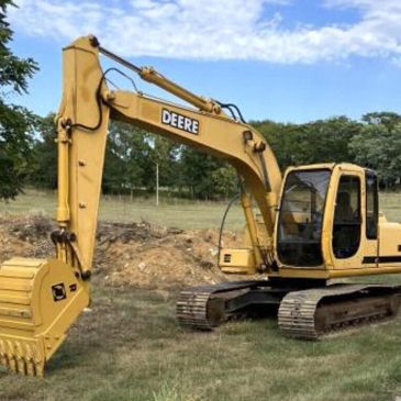 A yellow Deere excavator parked on grassy land with trees in the background.