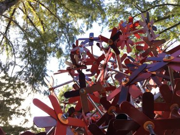 Colorful metal sculpture with flower shapes under tall trees in sunlight.