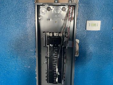 Open electrical panel with wires against a blue wall.