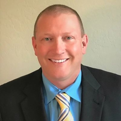 Smiling man in suit with blue shirt and striped tie against plain wall.