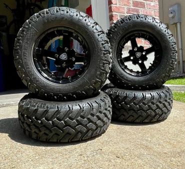 Four rugged off-road tires stacked in two pairs on a driveway.