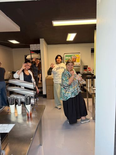 Four people smiling and waving inside a café or kitchen area.