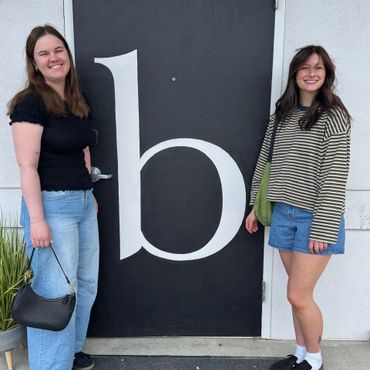 Two women smiling by a black door with a large white letter 'b'.