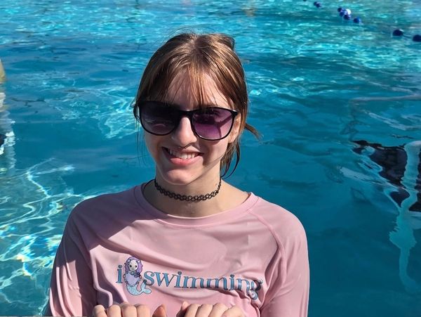 A smiling girl wearing sunglasses and a pink swimming shirt in a pool.