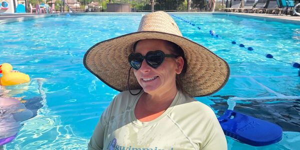 Woman in heart-shaped sunglasses and large hat smiles in a sunny pool.