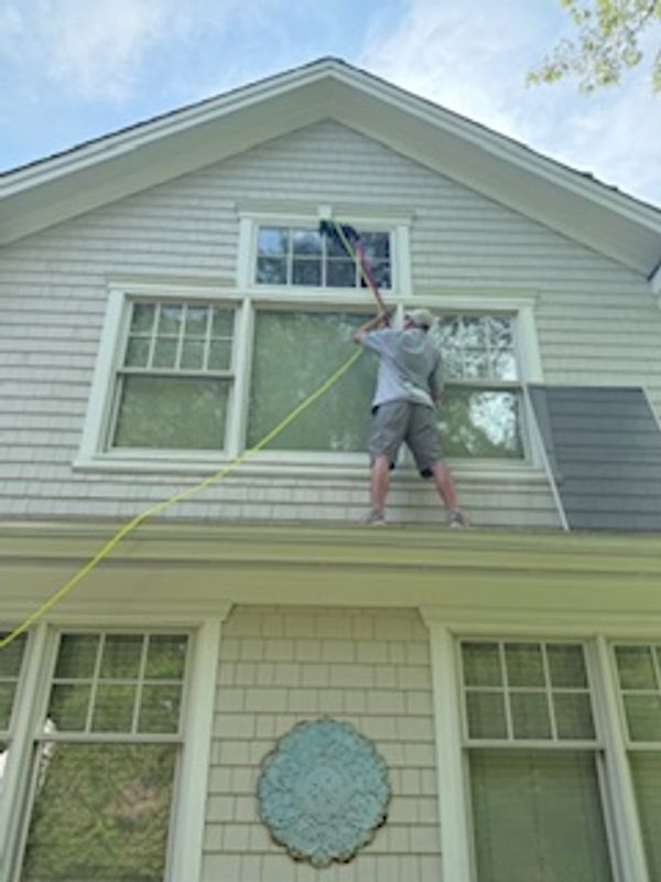 Man cleaning second-story window while standing on roof's edge.