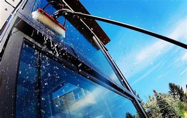 Cleaning a glass window with a brush on a long pole under clear blue sky.