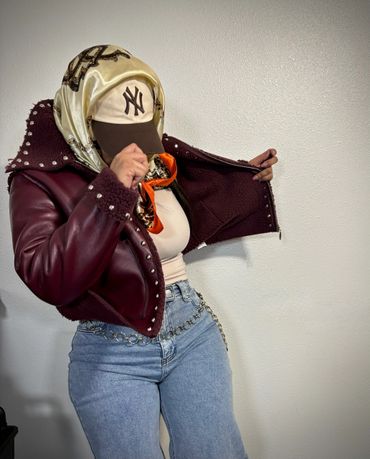 Person wearing a studded maroon jacket and a New York Yankees cap, posing against a plain wall.