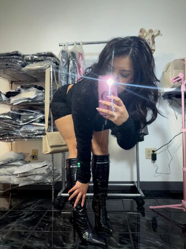 Woman in black outfit and knee-high boots taking a mirror selfie.