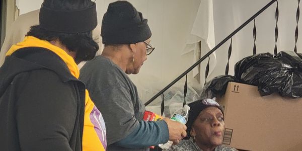 Three women engaged in conversation indoors near stairs with household items.
