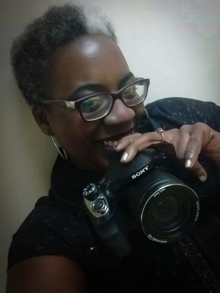 A photo of Gail a dark skin Black woman,  with a short black and grey afro. smiling with her camera 