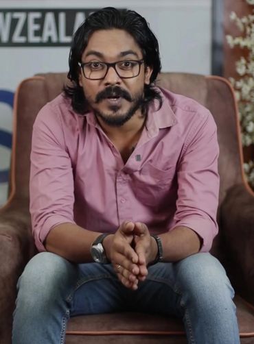 Man with glasses and beard sitting on a chair, speaking.