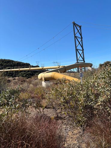 Large yellow pipeline supported by a metal structure in a dry, shrub-filled landscape under a clear blue sky.
