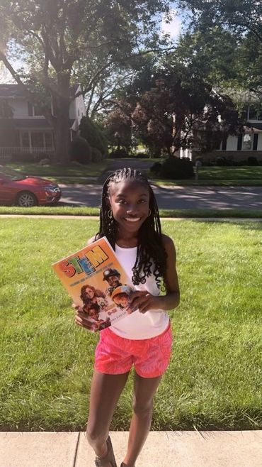 A young Black Girl with a white shirt and pink shorts is outside holding up our book