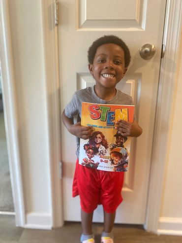A young Black Boy with short hair wears a grey shirt and red shorts, holding up our book