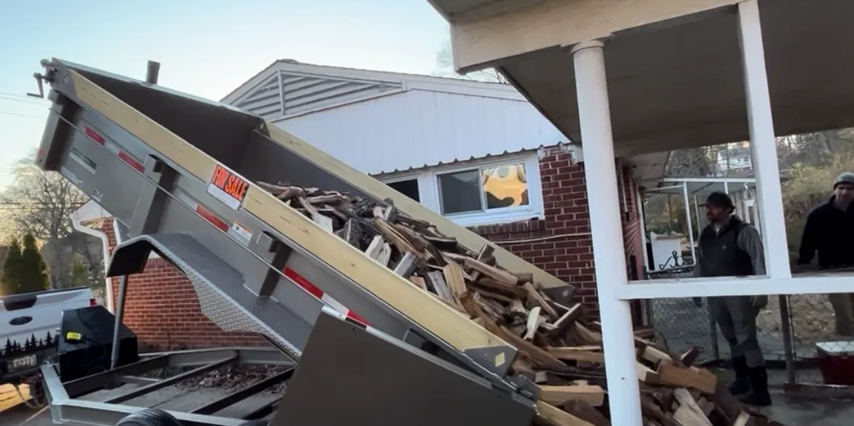 Truck dumping firewood next to porch with two men watching.