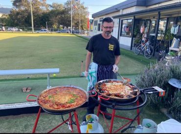 Man cooking two large pans of paella outdoors.