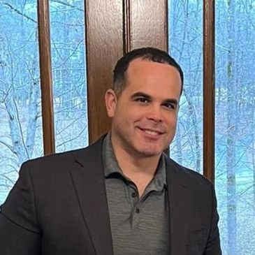 Man smiling in a dark blazer and gray shirt by a window.