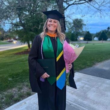 Woman smiling in graduation gown holding diploma and flowers outdoors.