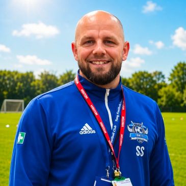 A smiling man in a blue sports jacket with a lanyard outdoors.