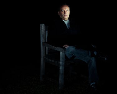 Man sitting on a wooden chair in dim lighting with a serious expression.