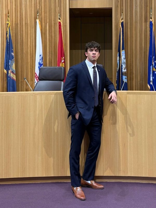 A young man in a suit stands confidently in a courtroom with flags behind him.