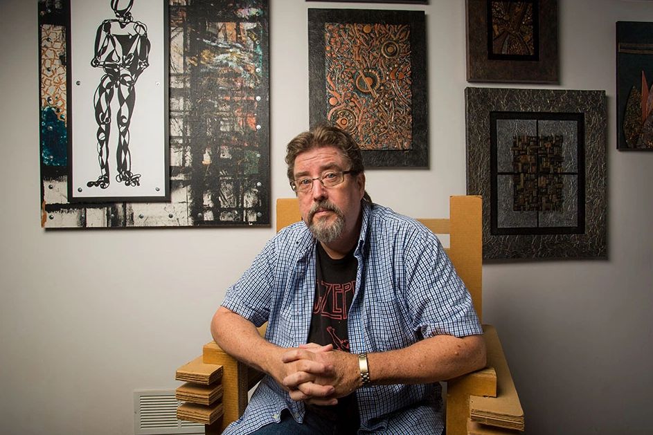 Dale white sitting on his cardboard chair with a collection of his work in the background. 