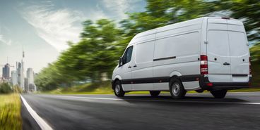 A white cargo van driving fast on a road with greenery and cityscape in the background.