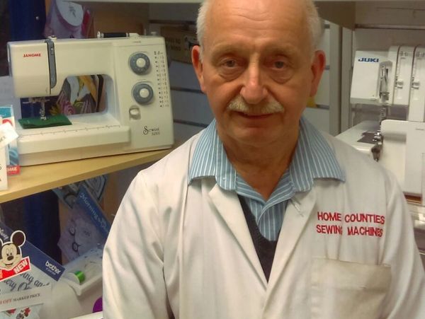 An older man in a white coat stands in front of sewing machines.