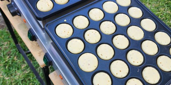 Mini pancakes cooking in a tray on an outdoor electric griddle.