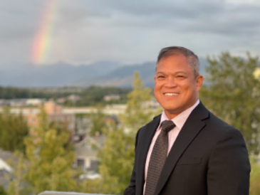 Smiling man in a suit with a rainbow and cityscape background.