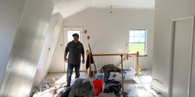 Two men working on renovating a bright, white attic room with tools and supplies scattered.