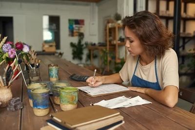 Woman writing at a wooden table covered with ceramics and notebooks.