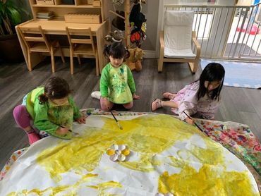 Three children painting a large yellow artwork on the floor indoors.