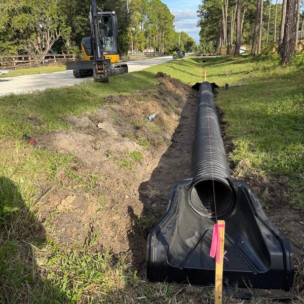 Drainage pipe being installed along a roadside ditch with excavation equipment nearby.