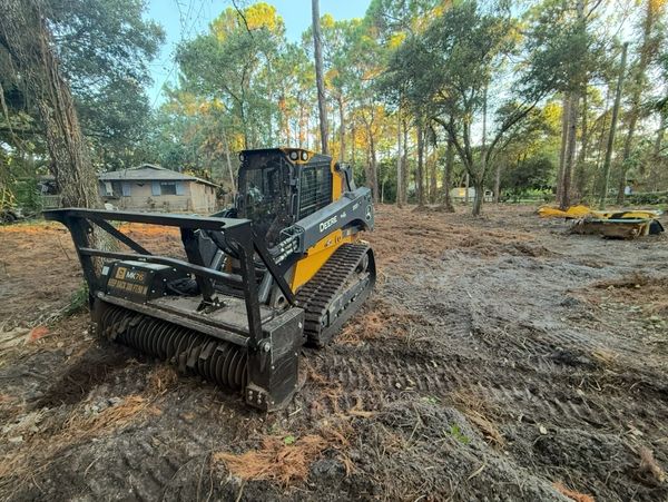 A John Deere compact track loader with forestry equipment clearing a wooded area.