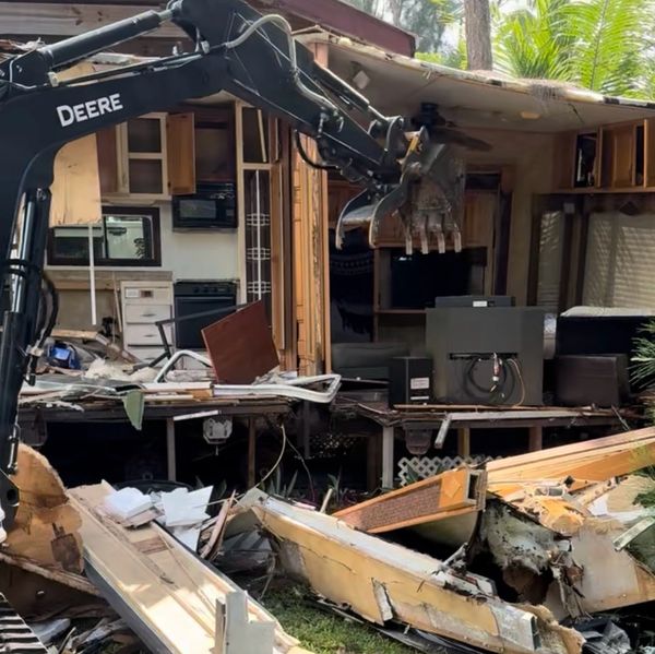 Excavator demolishing a wooden house, debris scattered around.