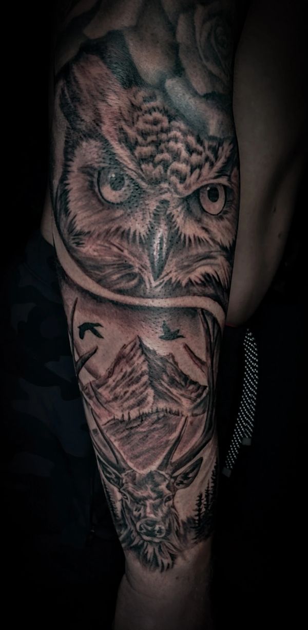 Detailed black and gray tattoo of an owl, mountain, and deer on an arm.