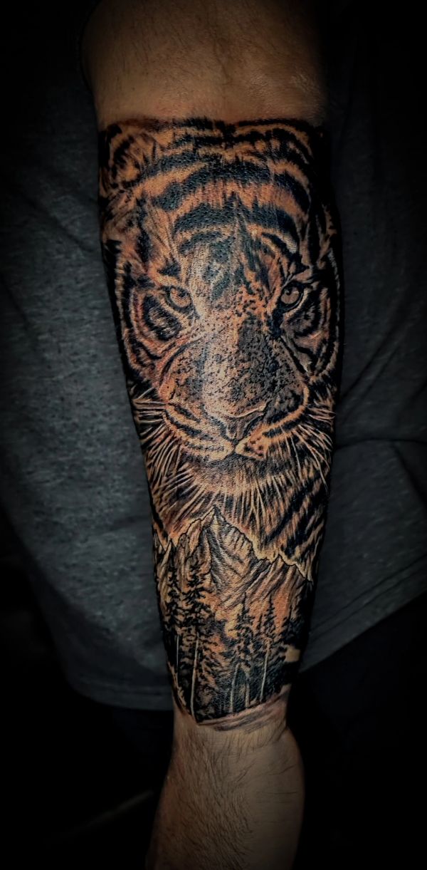 Tattoo of a fierce tiger face with a mountainous forest scene below it on a forearm.
