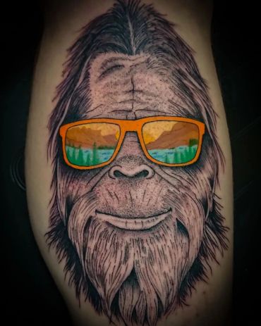 Tattoo of a smiling Bigfoot wearing orange sunglasses reflecting a nature scene.