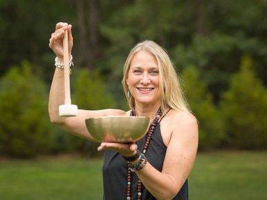 Woman holding a singing bowl and mallet outdoors, smiling.