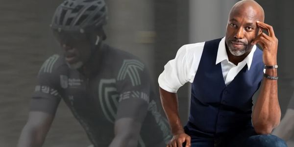 Marcus Allen in a vest, with his finger on his head, in front of a picture of him cycling.