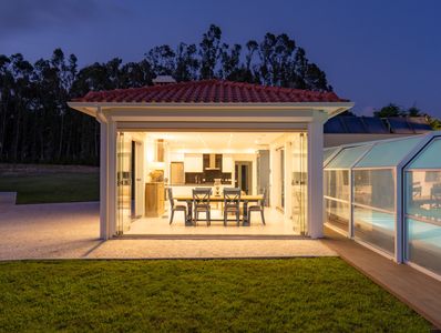 Modern outdoor kitchen and dining area with glass walls and poolside view.