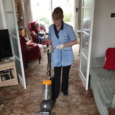 Caregiver vacuuming carpet while elderly person sits in background.