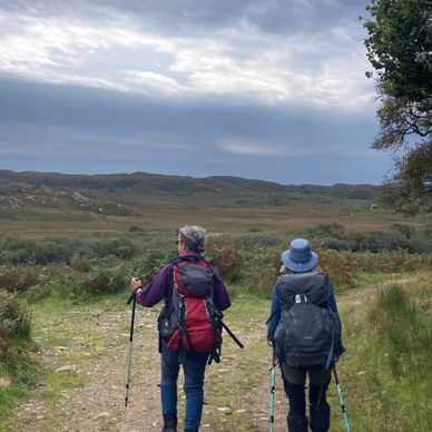 Two hikers walking on a trail in an open countryside under a cloudy sky.