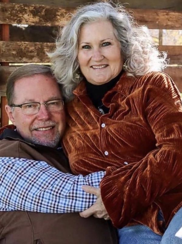 Smiling couple embracing outdoors with rustic wooden backdrop.