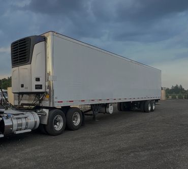 A large white refrigerated semi-trailer truck parked on a gravel lot under a cloudy sky.
