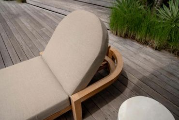 Beige cushioned wooden lounge chair on a wooden deck with greenery.